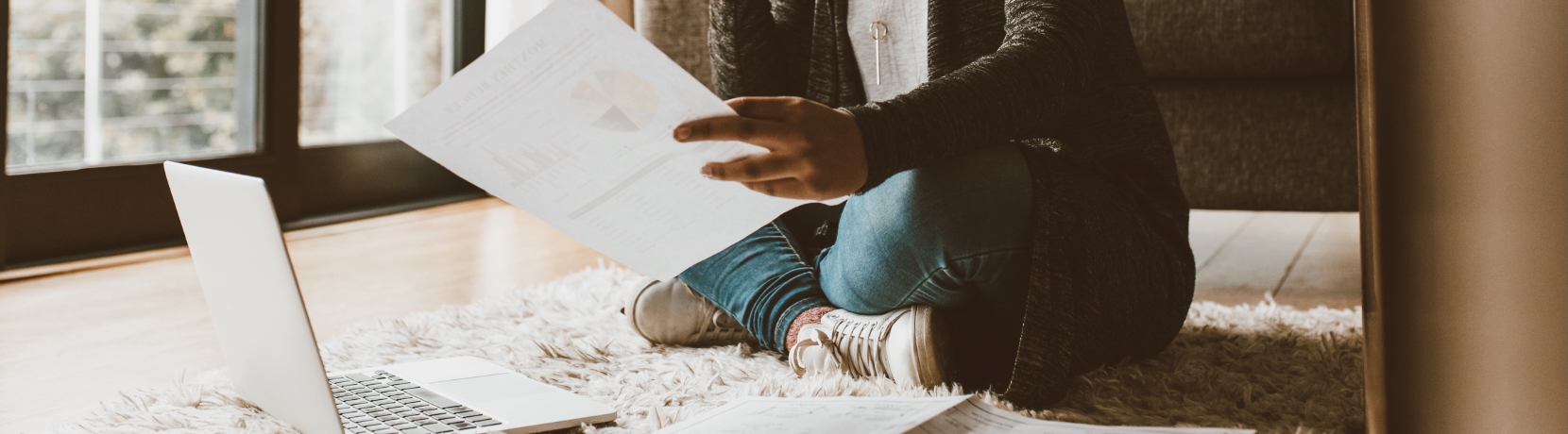 Woman sitting on the floor on her laptop looking at a piece of paper.