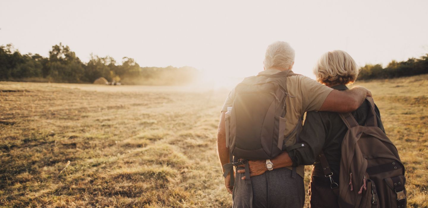An older couple with their backs to the camera standing in a field with backpacks on and their arms around one another.