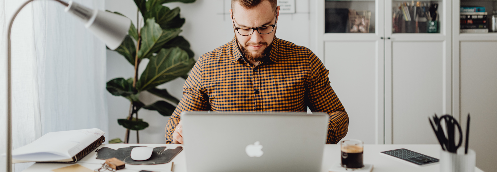 An image of a man sitting at a desk wearing glasses and looking at a laptop.