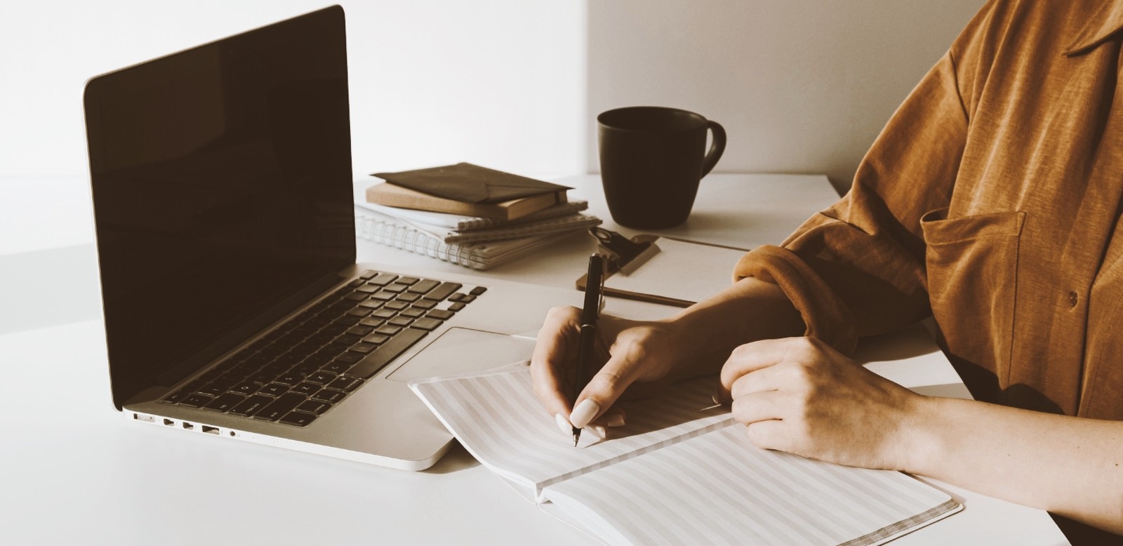 Person writing in a notebook with a laptop, books, and coffee mug sitting on the desk.